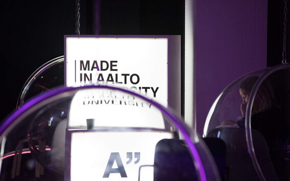 Woman sitting in a clear bubble chair beside a bright sign reading “Made in Aalto University” in a dark room
