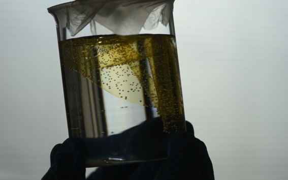 Gloved hands hold a clear beaker of yellow liquid with bubbles and a white filter paper on top