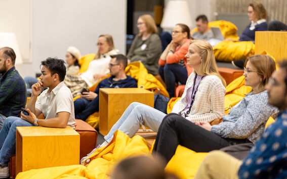 Participants in the Inventors programme sit on yellow beanbags and wooden blocks in a modern room, listening to a talk in the AGrid building at Aalto campus. 