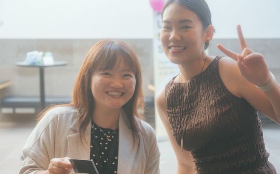 Two smiling women indoors, one in a light jacket holding a card, the other in a brown shirt making a peace sign