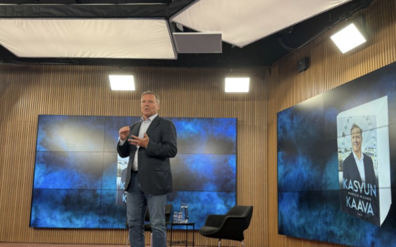Man in blazer and jeans speaks on stage with blue screens and book cover backdrop in studio setting