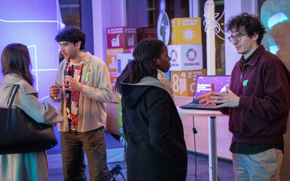 Four people talk at a tech event, two by a laptop, colourful UN goal posters glowing in the background