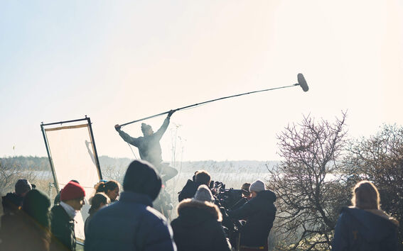 Outdoor film crew in winter clothes, boom mic raised high above a busy set near bare trees