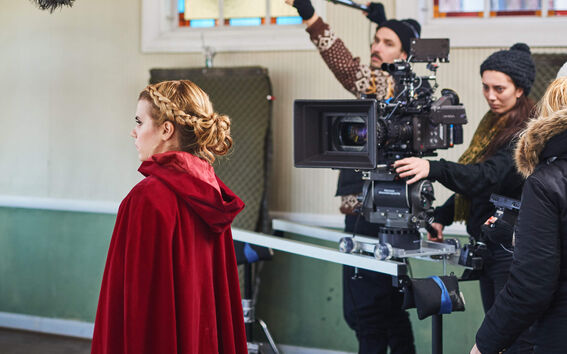 A woman in a red cape, with a camera crew in the background.