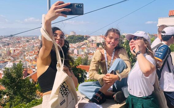 Three young people take a selfie on a sunny city viewpoint, with rooftops and trees stretching behind them.