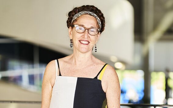 Smiling woman with glasses in patterned dress and headband, standing indoors in soft light