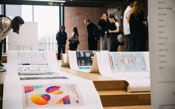 People in a hall viewing large infographic posters spread across wooden steps by a brick wall.