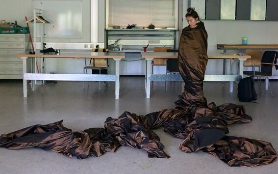 A person wrapped in brown fabric stands in a studio while the fabric spreads in folds across the floor.