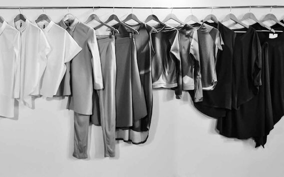 Row of minimalist shirts, tunics and trousers on hangers, fading from light to dark tones against a plain wall
