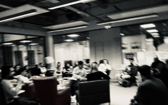 Blurry black‑and‑white photo of people sitting in a circle of chairs in an office‑style room