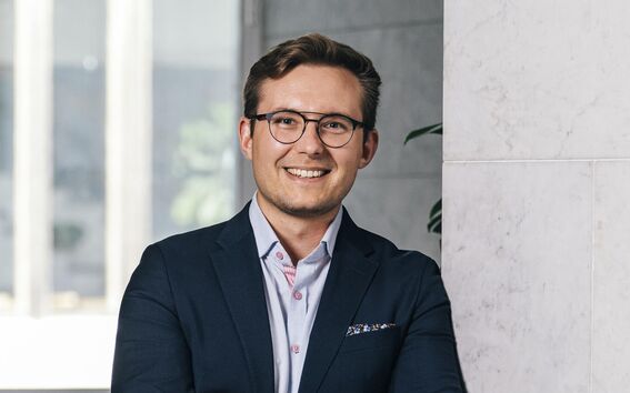 Smiling man in glasses and navy blazer, arms crossed, leaning against a light marble wall indoors
