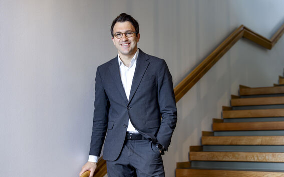 Smiling man in a dark suit and glasses stands by a wooden staircase in a bright hallway