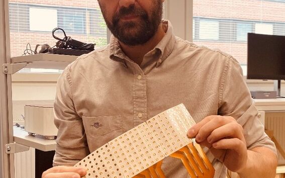 Man in beige shirt indoors holding perforated white and orange flexible circuit board with three connectors
