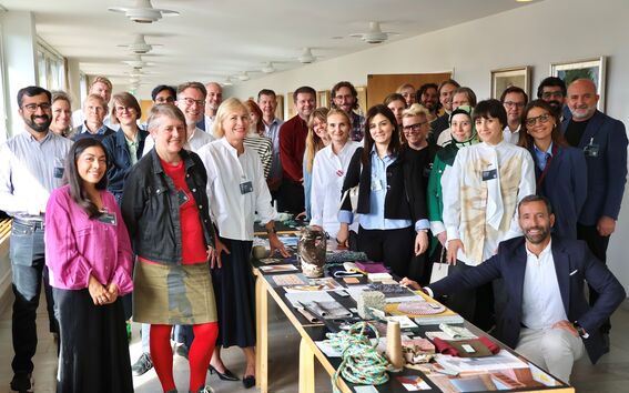 Large group indoors around a long table displaying craft materials and samples, smiling at the camera