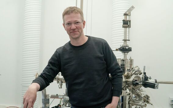 Mies tummassa paidassa seisoo valoisassa laboratoriossa metallilaitteen vieressä
