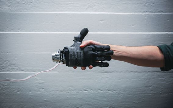 Human arm holding a black robotic hand with wires, against a light grey wall background