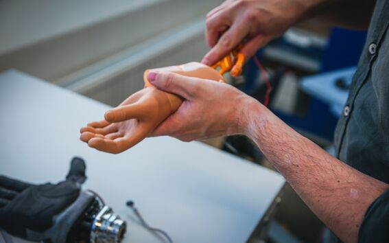 Person fitting a tan prosthetic hand onto an orange robotic arm at a workbench
