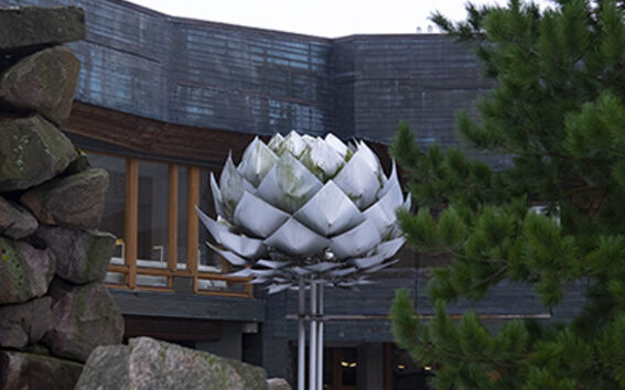 Metal lotus sculpture among rocks and snow outside a modern wooden and stone building with pine tree