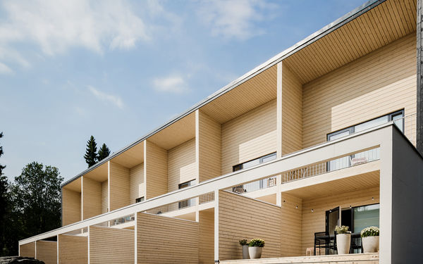 Modern row house with wooden cladding, large terraces, and potted plants outside, under a partly cloudy sky.