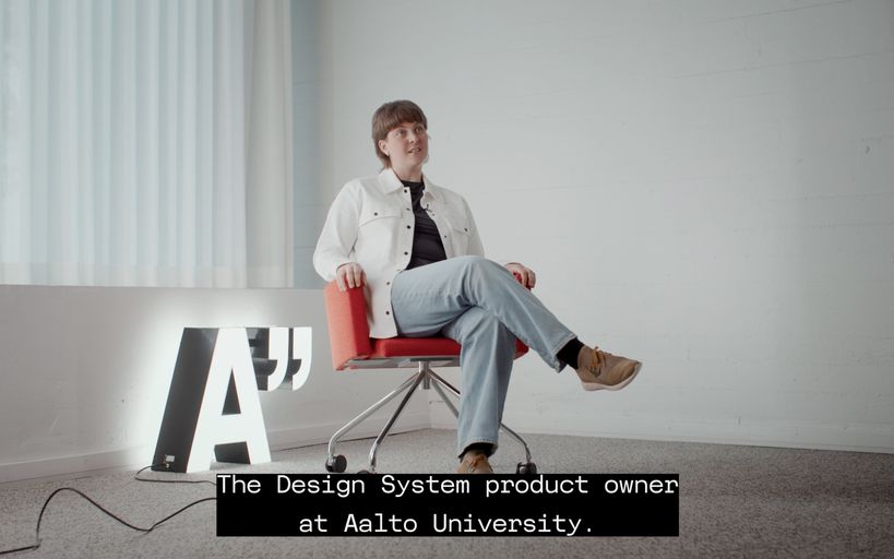 A woman sitting in a chair in a light and empty room, a lit A" logo on the floor behind her