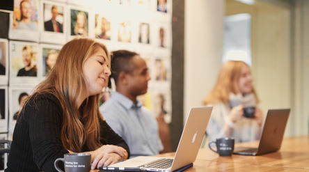 Three people sitting in front of the desk with laptops and coffee mugs. 