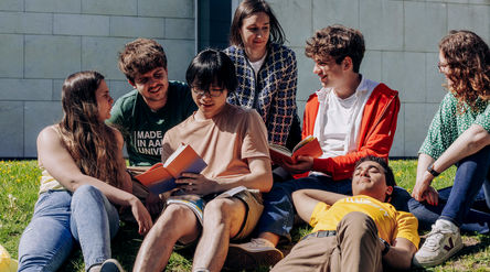 a group of students sitting and laying on the grass, two are reading a book. 