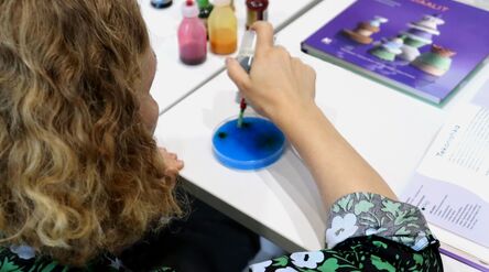 Nainen tiputtelee elintarvikeväriä tippapullosta petrimaljalle selkä katsojaan päin. Taustalla elintarvikevärejä ja Mainiot materiaalit kirja