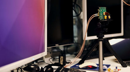Three monitors on a desk. A camera on a tripod focuses on a circuit board.