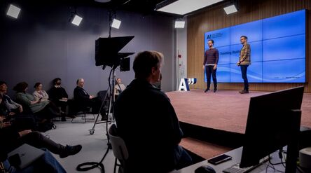 Two men stand on a stage presenting to a seated audience. The background screen displays 'Teaching Studio' with the Aalto University logo.