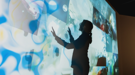 A person interacts with a large screen displaying various abstract images. The person’s silhouette is visible.