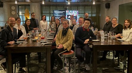 A group of people seated at a long table in a restaurant, drinking and dining, with a presentation screen in the background.