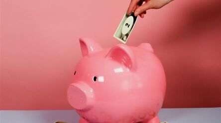 Tatuoitu käsi asettaa setelin vaaleanpunaiseen säästöpossuun. Useita seteleitä sen ympärillä.