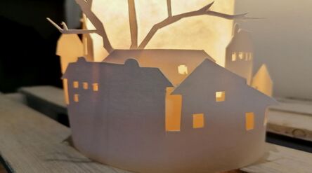 Paperilyhty kuvaa taloja ja puita leikattuina kuvioina, lämmin valo valaisee sisältä.