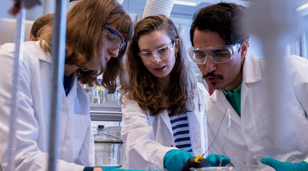 Three people wearing lab coats and teal gloves conducting an experiment in a laboratory.