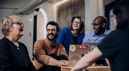 Viisi ihmistä kokoontuneena läppärin ympärille rento toimistoympäristössä, teollinen sisustus.