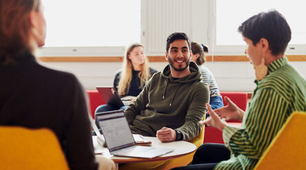 Joukko ihmisiä keskustelemassa pöydän ympärillä kannettavien tietokoneiden kanssa, istuen värikkäillä tuoleilla valoisassa huoneessa.