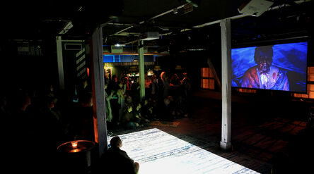 Hämärästi valaistu yökerhon tapahtumatila, jossa valkokankaalla näytetään elokuvaa ja lattialla aaltoilee yleisön 
katsellessa.