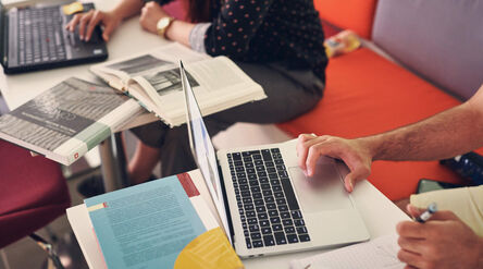 Two people studying at a table with laptops and books. One person is typing, the other is writing.