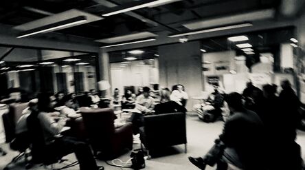 A blurred image of a group of people sitting and discussing in a modern office space with glass walls.