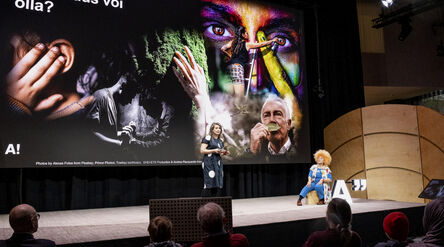 Esitys, jossa suuri näyttö näyttää värikkäitä kuvia ja tekstiä. Kaksi ihmistä lavalla, yksi seisoo ja yksi istuu.