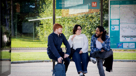 Three students at bus stop, with a map board and greenery in the background.