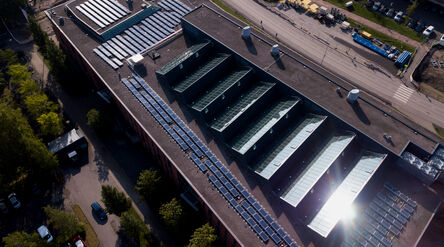 Aerial view of large flat-roof building with glass skylights, solar panels and nearby road with parked vehicles