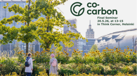 Two people in light coats walk through a sunny urban garden with tall trees and flowers, Helsinki buildings behind