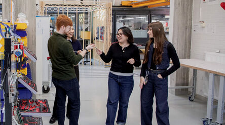 Four students talk in a workshop, surrounded by tools, workbenches and industrial equipment.