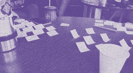 Workshop photo digitally modified with a purple duotone effect. The picture shows a table with sticky notes, a pen, a phone, a cup, thermos and mate, with people and other table in the background.