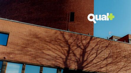 Red brick building with bare tree shadows and blue sky, white text logo “Qual+” in the top right
