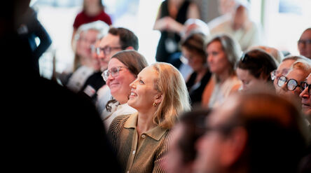 Smiling woman with blond hair in a brown shirt sits among an attentive indoor audience