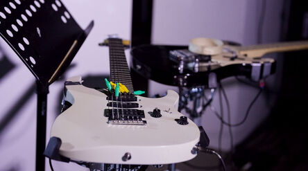 White electric guitar with colourful picks on strings, next to a black guitar and music stand on a dim stage.