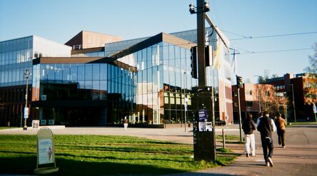 Modern glass-fronted building by a grassy street as three people walk past in bright daylight