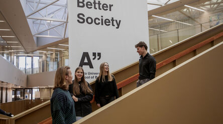 Four students chat on indoor stairs beside a large white Business–Better Society sign in a modern atrium